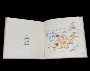 Image of open book against a black background. The left page has a short Hebrew poem. The right page has a full frame, hand-colored drawing featuring a small sailboat at sea, with fish visible below. Two of the fish, one orange and one blue, are larger than life and larger than the boat.