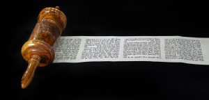 Photograph of partially unrolled scroll against a black background. The scroll is housed in an ornate wooden case. Four columns of text are visible on the unrolled paper.